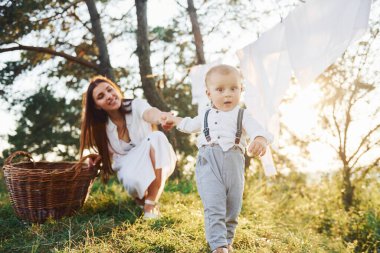 Kuruması için ipe asılmış temiz giysiler. Küçük oğluyla birlikte genç bir anne ormanda. Güzel güneş ışığı.