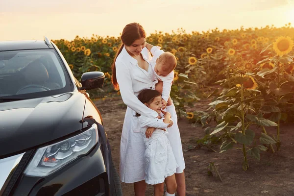 Near modern black car. Young mother with her little son and daughter is outdoors in the agricultural field. Beautiful sunshine.