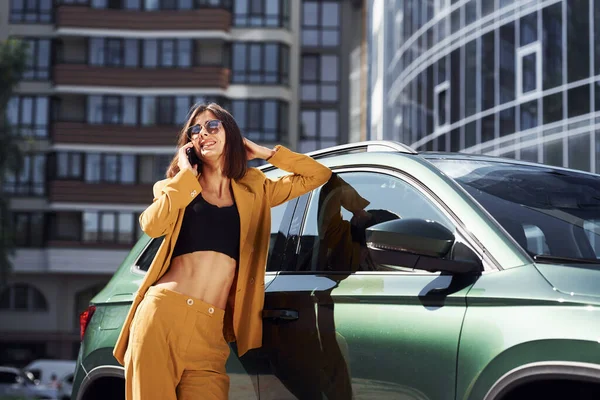 Talks by the phone. Young fashionable woman in burgundy colored coat at daytime with her car.
