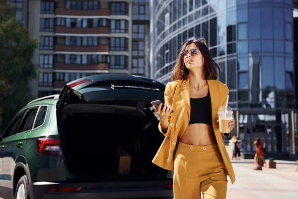 近代的なビジネスビルを背景に 若いですファッショナブルな女性でバーガンディ色のコートに昼間とともに彼女の車 — ストック写真