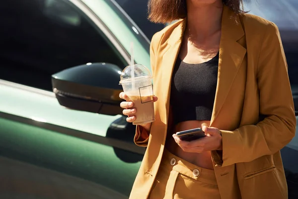 飲み物とスマートフォンを持っている 若いですファッショナブルな女性でバーガンディ色のコートに昼間とともに彼女の車 — ストック写真