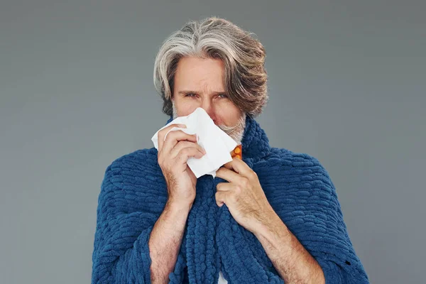 Getting sick. Against grey background. Stylish modern senior man with gray hair and beard is indoors.