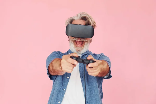 Playing game with virtual reality glasses and joystick. Stylish modern senior man with gray hair and beard is indoors.