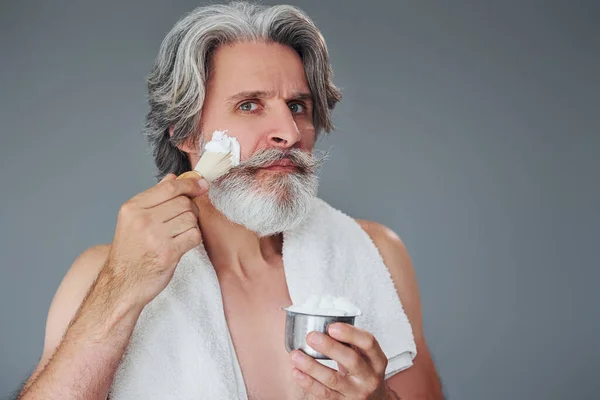 With shaving gel. Stylish modern senior man with gray hair and beard is indoors.