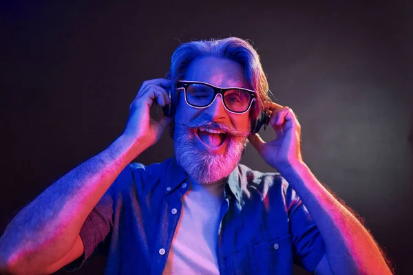 Neon lighting. In headphones. Stylish modern senior man with gray hair and beard is indoors.
