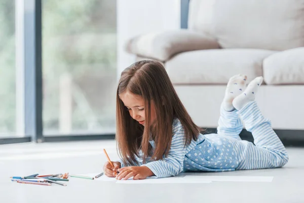 Lying on the floor and drawing. Cute little girl indoors at home alone.