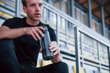 Bir şişe suyla oturuyor. Gündüz vakti siyah gömlekli ve pantolonlu genç bir adam..