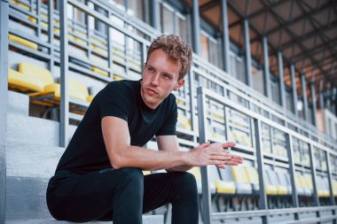 Merdivenlerde oturuyor. Gündüz vakti siyah gömlekli ve pantolonlu genç bir adam..