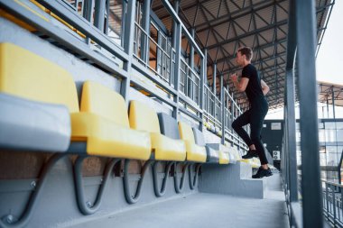 Tribünde koşuyor. Gündüz vakti siyah gömlekli ve pantolonlu genç bir adam..