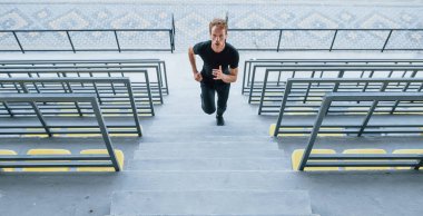 Tribünde koşuyor. Gündüz vakti siyah gömlekli ve pantolonlu genç bir adam..