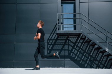 Siyah binanın yakınında koşuyor. Gündüz vakti siyah gömlekli ve pantolonlu genç bir adam..