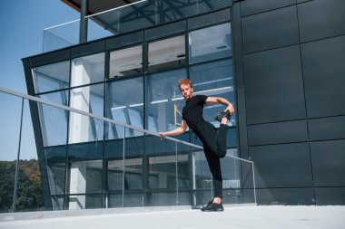 Siyah duvarlı binanın yakınında egzersiz yapıyor. Gündüz vakti siyah gömlekli ve pantolonlu genç bir adam..