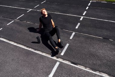 Yorgun koşucu rayına oturur ve mola verir. Gündüz vakti siyah gömlekli ve pantolonlu genç bir adam..