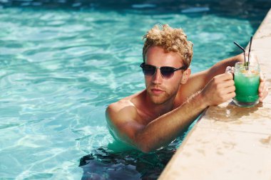 Siyah güneş gözlüklü ve kokteylli adam yüzme havuzunda.