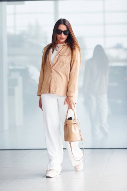 Zarif giyinmiş ve güneş gözlüklü bir kadın yansıtıcı arka plana karşı içeride duruyor..