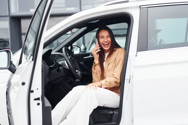 Talks by phone with door opened. Fashionable beautiful young woman and her modern automobile.
