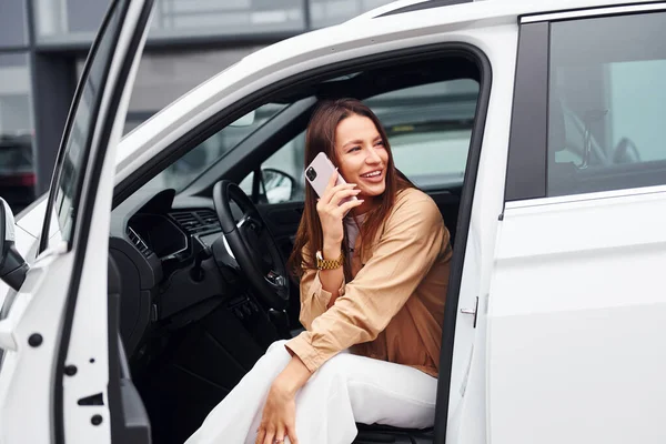 Talks by phone with door opened. Fashionable beautiful young woman and her modern automobile.