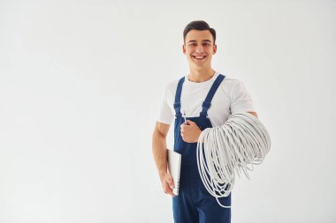 Kablolar, not defteri ve tornavida tutuyor. Mavi üniformalı erkek işçi beyaz arka planda stüdyonun içinde duruyor..