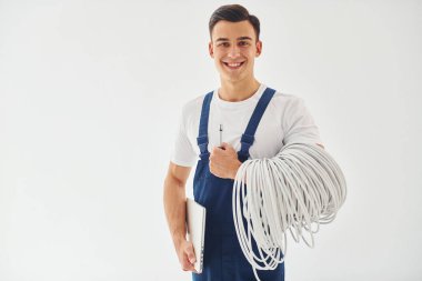 Kablolar, not defteri ve tornavida tutuyor. Mavi üniformalı erkek işçi beyaz arka planda stüdyonun içinde duruyor..