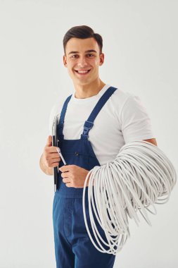 Kablolar, not defteri ve tornavida tutuyor. Mavi üniformalı erkek işçi beyaz arka planda stüdyonun içinde duruyor..