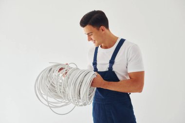 Kablo tutuyor. Mavi üniformalı erkek işçi beyaz arka planda stüdyonun içinde duruyor..
