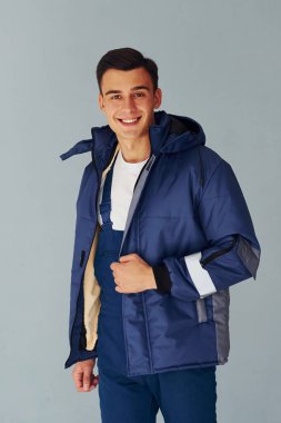 Kalın ceketli. Mavi üniformalı erkek işçi beyaz arka planda stüdyonun içinde duruyor..