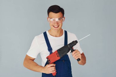 Koruyucu gözlüklerle ve ellerinde matkapla. Mavi üniformalı erkek işçi beyaz arka planda stüdyonun içinde duruyor..