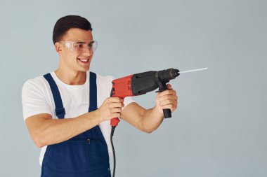 Koruyucu gözlüklerle ve ellerinde matkapla. Mavi üniformalı erkek işçi beyaz arka planda stüdyonun içinde duruyor..