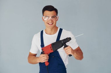 Elleri matkapla delin. Mavi üniformalı erkek işçi beyaz arka planda stüdyonun içinde duruyor..