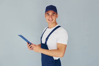 Şapka ve not defteriyle. Mavi üniformalı erkek işçi beyaz arka planda stüdyonun içinde duruyor..