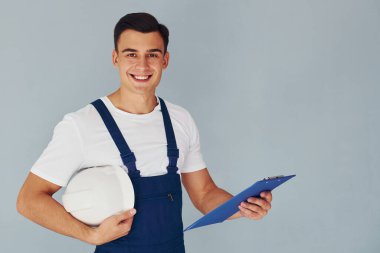 Not defteri ve kasketli. Mavi üniformalı erkek işçi beyaz arka planda stüdyonun içinde duruyor..