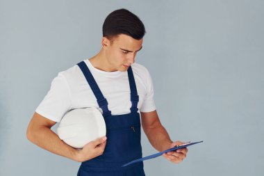 Not defteri ve kasketli. Mavi üniformalı erkek işçi beyaz arka planda stüdyonun içinde duruyor..