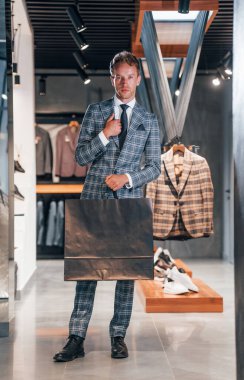 Çantayı satışta tutuyor. Modern mağazada yeni kıyafetleri olan genç bir adam. Erkekler için pahalı bir elbise..