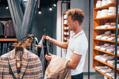 Ceket arıyor. Modern mağazada yeni kıyafetleri olan genç bir adam. Erkekler için pahalı bir elbise..