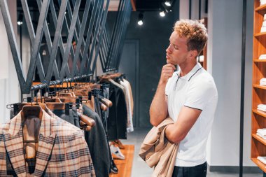 Ceket arıyor. Modern mağazada yeni kıyafetleri olan genç bir adam. Erkekler için pahalı bir elbise..