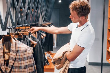 Ceket arıyor. Modern mağazada yeni kıyafetleri olan genç bir adam. Erkekler için pahalı bir elbise..