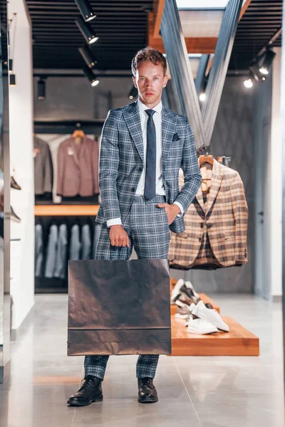 Çantayı satışta tutuyor. Modern mağazada yeni kıyafetleri olan genç bir adam. Erkekler için pahalı bir elbise..