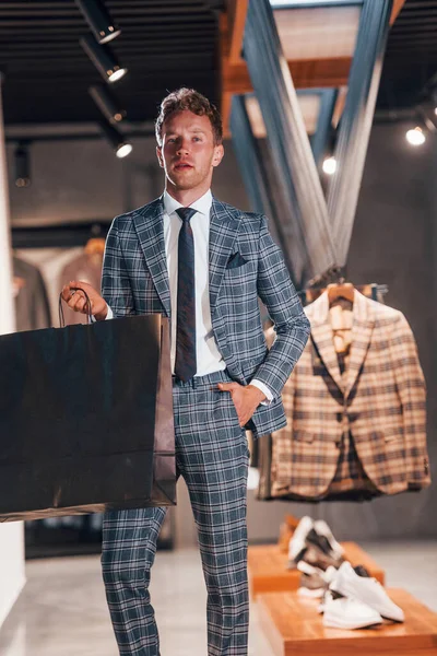 Çantayı satışta tutuyor. Modern mağazada yeni kıyafetleri olan genç bir adam. Erkekler için pahalı bir elbise..