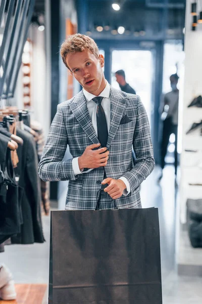 Çantayı satışta tutuyor. Modern mağazada yeni kıyafetleri olan genç bir adam. Erkekler için pahalı bir elbise..