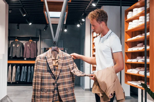 Ceket arıyor. Modern mağazada yeni kıyafetleri olan genç bir adam. Erkekler için pahalı bir elbise..