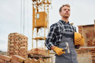 Ayakta duruyor ve çekiçle kaskı tutuyor. Üniformalı inşaat işçisi ve güvenlik ekipmanı inşaatında çalışıyor.