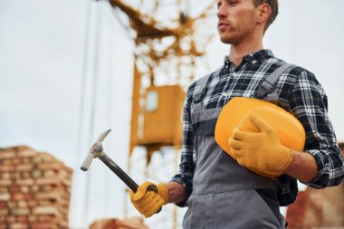 Ayakta duruyor ve çekiçle kaskı tutuyor. Üniformalı inşaat işçisi ve güvenlik ekipmanı inşaatında çalışıyor.