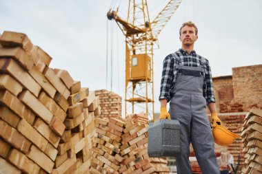 Kasket ve çanta ellerinde. Üniformalı inşaat işçisi ve güvenlik ekipmanı inşaatında çalışıyor.