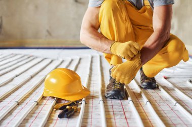 Kasketli. Sarı üniformalı işçiler zemin altı ısıtma sistemi kuruyor.