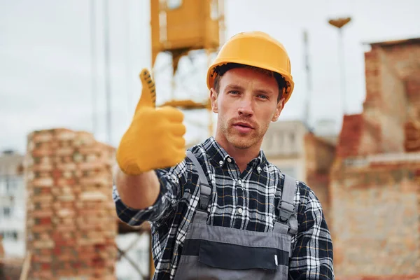 Shows thumb up. Construction worker in uniform and safety equipment have job on building.