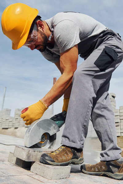 Uses circular saw. Male worker in yellow colored uniform have job with pavement.