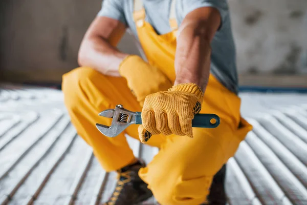 Holds wrench in hand. Worker in yellow colored uniform installing underfloor heating system.