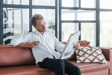 Gazete okuyor. Beyaz, zarif beyaz gömlekli beyaz adam evde..