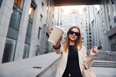 Bir bardak içki tutuyor. Güzel genç Avrupalı kadın gündüz vakti açık havada şehirde..