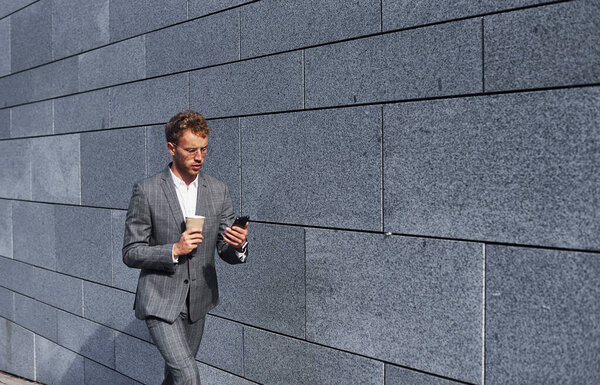 With phone and drink in hands. Young successful businessman in grey formal wear is outdoors in the city.